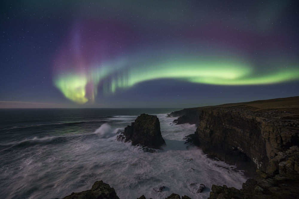 1500px by 500px image of an Aurora filled with Purple, Green, and Pink over a wild Atlantic sea that is crashing into Scottish cliffs 